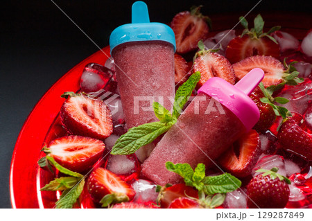 Two trawberry ice cream popsicles on red plate with ice cubes chopped strawberries and mint leaves on a black background. Strawberry ice cream popsicles. Horizontal. Two trawberry ice cream popsicles on red plate with ice cubes chopped strawberries and mint leaves on a black background. Strawberry ice cream popsicles. Horizontal. 129287849
