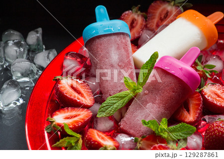 Colorful strawberry ice cream and popsicles rest in a vibrant bowl filled with crushed ice and fresh strawberries, creating a perfect summer dessert. The scene invites enjoyment. 129287861