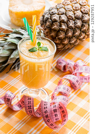 Diagonally left down a glass of pineapple smoothie with straws and mint leaf and open centimeter in the background pineapple and slice, on yellow checkered tablecloth. Close. Vertical. Diagonally left down a glass of pineapple smoothie with straws and mint leaf and open centimeter in the background pineapple and slice, on yellow checkered tablecloth. Close. Vertical. 129287951