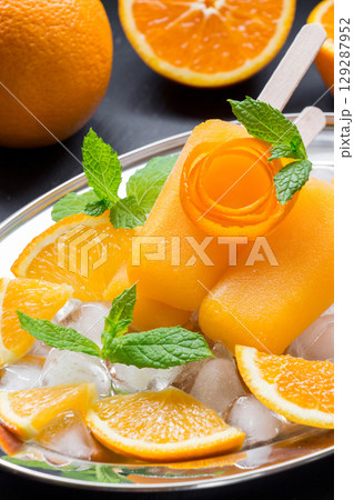 Orange sorbet ice cream decorated with a flower of orange peel on silver plate with ice cubes, orange pieces, mint near orange slices on black background. Orange sorbet ice cream popsicles. Vertical. Orange sorbet ice cream decorated with a flower of orange peel on silver plate with ice cubes, orange pieces, mint near orange slices on black background. Orange sorbet ice cream popsicles. Vertical. 129287952