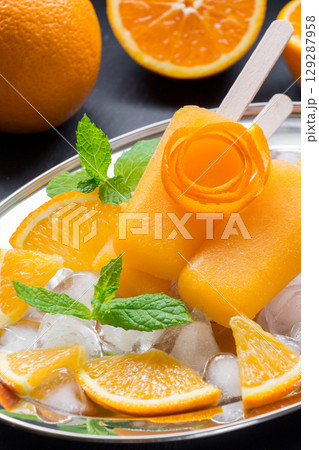 Orange sorbet ice cream decorated with a flower of orange peel on silver plate with ice cubes, orange pieces, mint near orange slices on black background. Orange sorbet ice cream popsicles. Vertical. 129287958