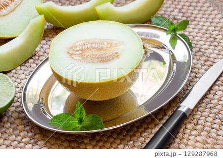 Diagonally at the center on a silver platter half of melon near melon slices, mint leaves, knife on a straw napkin background. Half of melon on silver platter. Horizontal. Close. Diagonally at the center on a silver platter half of melon near melon slices, mint leaves, knife on a straw napkin background. Half of melon on silver platter. Horizontal. Close. 129287968
