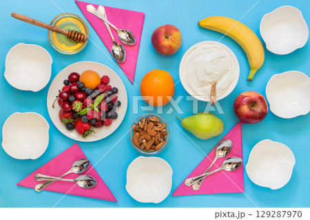 Greek yogurt around a variety of fruits and berries, nuts, honey and 6 empty bowls, 6 spoons and napkins on a light blue background. Yogurt and a variety of ingredients for it. Horizontal. Top view. Greek yogurt around a variety of fruits and berries, nuts, honey and 6 empty bowls, 6 spoons and napkins on a light blue background. Yogurt and a variety of ingredients for it. Horizontal. Top view. 129287970