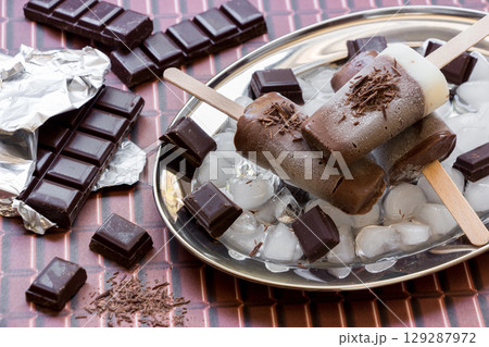 Chocolate vanilla ice cream on a stick on a silver platter with ice and pieces of chocolate near pieces of chocolate on a chocolate background. Chocolate vanilla ice cream popsicles. Horizontal. Chocolate vanilla ice cream on a stick on a silver platter with ice and pieces of chocolate near pieces of chocolate on a chocolate background. Chocolate vanilla ice cream popsicles. Horizontal. 129287972