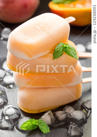 Three mango ice cream popsicles are piled on top of each other on a dark surface. They are surrounded by ice cubes, suggesting a refreshing treat perfect for a warm summer day. 129288001