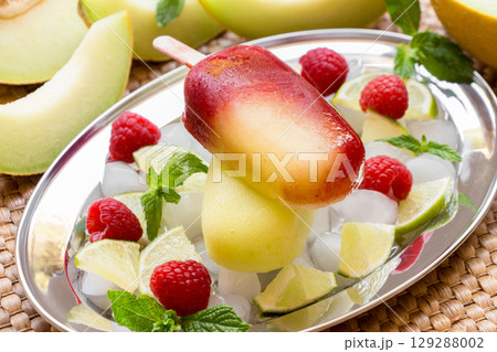 A vibrant melon raspberry ice cream sits atop a mound of ice cubes, surrounded by fresh raspberries, mint leaves, and sliced lime. 129288002