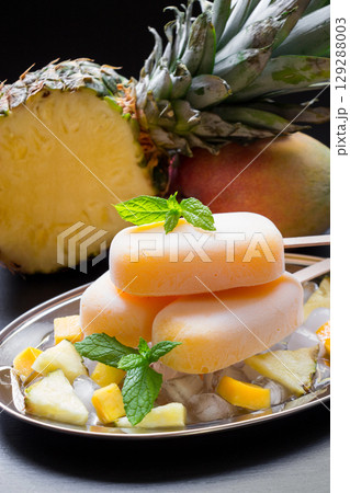 Exotic sorbet ice cream popsicles on silver plate with ice cubes chopped pineapple mango pieces, mint near half of pineapple, mango on a black background. Pineapple mango sorbet ice cream popsicles. Exotic sorbet ice cream popsicles on silver plate with ice cubes chopped pineapple mango pieces, mint near half of pineapple, mango on a black background. Pineapple mango sorbet ice cream popsicles. 129288003