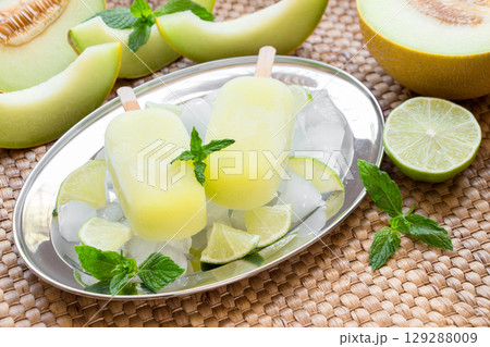 Enjoy refreshing melon ice cream popsicles presented on a silver tray surrounded by ice, mint, and lime, ideal for a warm afternoon gathering with friends or family. 129288009