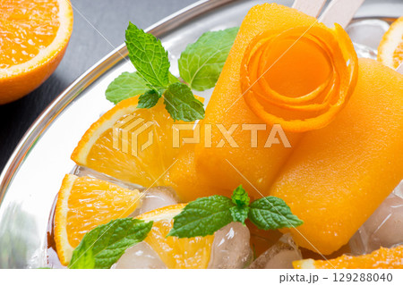 Orange sorbet ice cream decorated with a flower of orange peel, mint leaves on silver plate with ice cubes, orange slices on black background. Orange sorbet ice cream popsicles. Horizontal. Close. Orange sorbet ice cream decorated with a flower of orange peel, mint leaves on silver plate with ice cubes, orange slices on black background. Orange sorbet ice cream popsicles. Horizontal. Close. 129288040