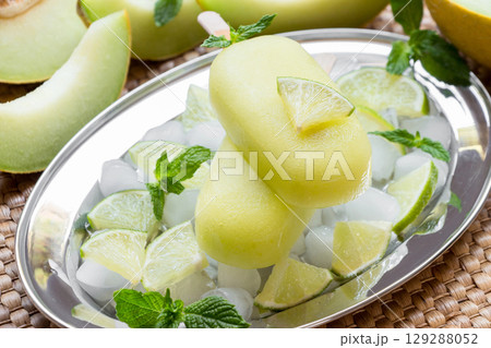 Melon and lime sorbet ice cream on silver plate with ice cubes, lime pieces, mint leaves near melon slices, lime on a straw napkin background. Melon and lime sorbet ice cream popsicles. Horizontal. Melon and lime sorbet ice cream on silver plate with ice cubes, lime pieces, mint leaves near melon slices, lime on a straw napkin background. Melon and lime sorbet ice cream popsicles. Horizontal. 129288052