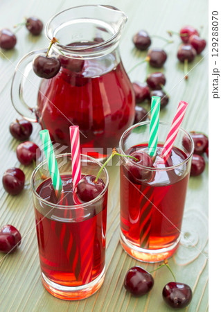 Two glasses of cherry juice with straws in the foreground and in the background a jar full of cherry juice. 2 cherry juices. Vertical. Close-up. Daylight. 129288070