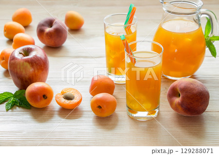 Right apricot peach apple juice in glasses with ice near the jar of juice and scattered apricots, peaches, apple, left empty space on light wooden background. Apricot peach apple juice with ice. 129288071
