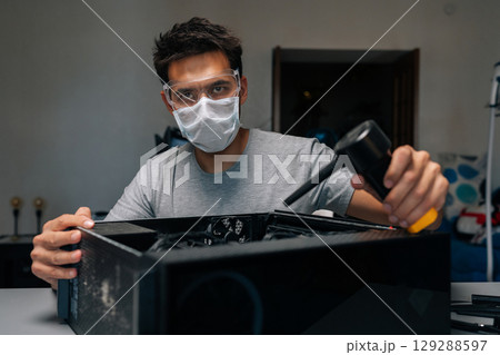Portrait of technician in face mask and safety glasses looking at camera cleaning computer case using small vacuum cleaner, demonstrating proper maintenance procedures. Concept of service computer. 129288597