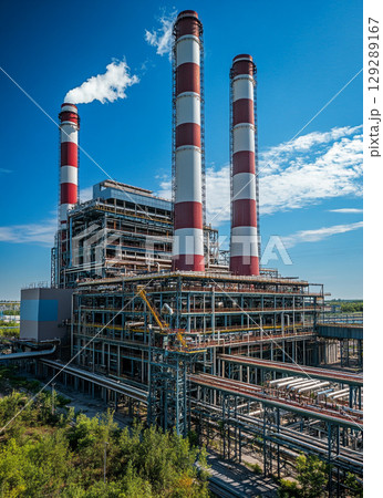 A large power plant with four red and white chimney towers stands against the blue sky, surrounded by lush green vegetation. 129289167