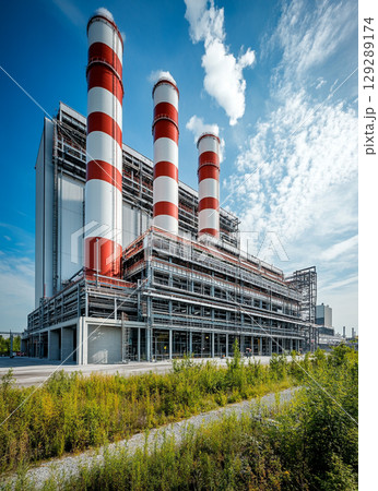 A large power plant with four red and white chimney towers stands against the blue sky, surrounded by lush green vegetation. 129289174
