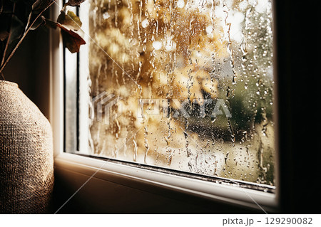 Warm autumn colors fill the view outside the window, where raindrops trickle down glass, creating a serene atmosphere. A vase adds a touch of nature to the indoor scene 129290082