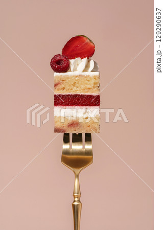 Vanilla sponge bento cake layered with raspberry and strawberry, topped with whipped cream and fresh fruit, presented on a golden fork against a soft pastel background, dessert concept 129290637
