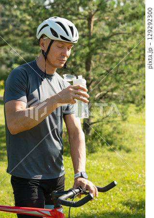 Male cyclist standing and near bike and drinking water at park. Summer. Vertical Male cyclist standing and near bike and drinking water at park. Summer. Vertical 129292300