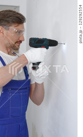 Professional male construction worker in a blue jumpsuit, safety goggles, and work gloves is screwing a screw into the wall using a cordless screwdriver, carrying out a careful home renovation Professional male construction worker in a blue jumpsuit, safety goggles, and work gloves is screwing a screw into the wall using a cordless screwdriver, carrying out a careful home renovation 129292554