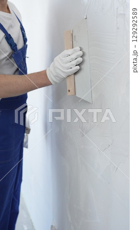 Male construction worker wearing protective gloves and blue construction coveralls, spreading plaster smoothly across wall using professional drywall taping knife during renovation, vertical view 129292589