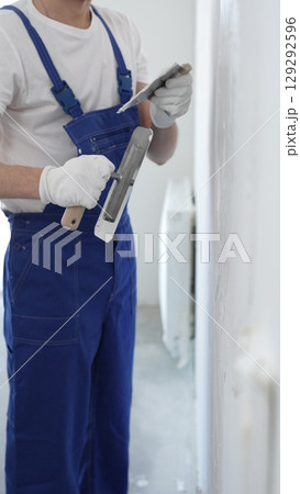 Professional construction worker wearing blue work overall is using a finishing trowel to carefully apply plaster on a wall, demonstrating expertise in home renovation 129292596