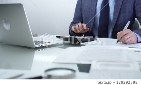 Businessman or accountant wearing blue suit is working with laptop, calculator and documents in office. Low lighting, close-up view. Business people, audit concept 129292598
