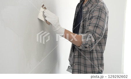 Unknown construction worker man wearing gloves and plaid shirt is using a putty knife to apply plaster on a white wall, during a home renovation or construction project 129292602