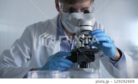 Male scientist researcher wearing blue gloves, protective glasses and medical mask, is using microscope in modern laboratory, portrait view. Low key lighting photo. Science and Medicine Male scientist researcher wearing blue gloves, protective glasses and medical mask, is using microscope in modern laboratory, portrait view. Low key lighting photo. Science and Medicine 129292609