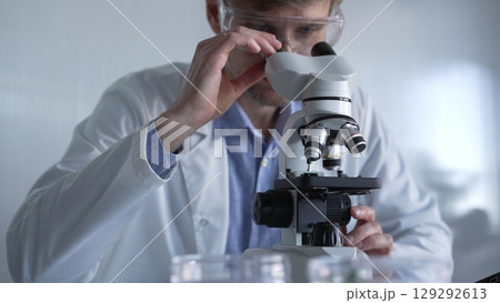 Male scientist wearing lab coat and protective glasses using microscope doing research in modern laboratory setup, portrait view. Low key lighting photo. Science and Medicine concept Male scientist wearing lab coat and protective glasses using microscope doing research in modern laboratory setup, portrait view. Low key lighting photo. Science and Medicine concept 129292613