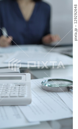 Businesswoman wearing blue dress is working with laptop, calculator and documents in a professional office setting. Low lighting, close-up vertical view. Business people, audit concept 129292648