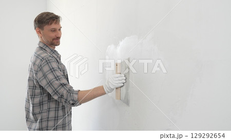 Man construction worker wearing protective gloves and plaid shirt, spreading plaster smoothly across wall surface using professional drywall taping knife during renovation project Man construction worker wearing protective gloves and plaid shirt, spreading plaster smoothly across wall surface using professional drywall taping knife during renovation project 129292654