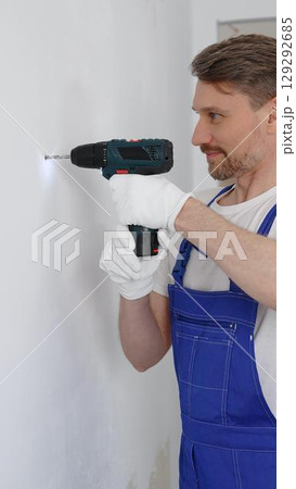 Man construction worker in a blue jumpsuit, safety goggles, and work gloves is screwing a screw into the wall using a cordless screwdriver, carrying out a careful home renovation. Vertical portrait Man construction worker in a blue jumpsuit, safety goggles, and work gloves is screwing a screw into the wall using a cordless screwdriver, carrying out a careful home renovation. Vertical portrait 129292685