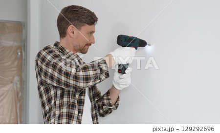 Man construction worker in checked shirt and safety gloves is screwing a screw into the wall using a cordless screwdriver, carrying out a careful home renovation. Horizontal portrait view Man construction worker in checked shirt and safety gloves is screwing a screw into the wall using a cordless screwdriver, carrying out a careful home renovation. Horizontal portrait view 129292696