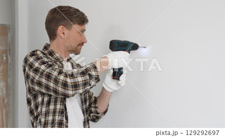 Middle aged man wearing checked shirt and white gloves, is drilling hole in a wall with cordless drill during home renovation work. Portrait view Middle aged man wearing checked shirt and white gloves, is drilling hole in a wall with cordless drill during home renovation work. Portrait view 129292697
