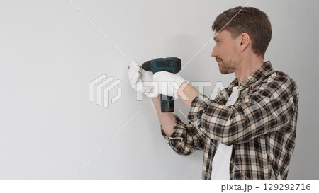 Middle aged man wearing checked shirt and white gloves, is drilling hole in a wall with cordless drill during home renovation work. Portrait view Middle aged man wearing checked shirt and white gloves, is drilling hole in a wall with cordless drill during home renovation work. Portrait view 129292716