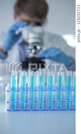 Test tubes with a blue liquid, close-up view. Male scientist wearing blue protective gloves and glasses is using microscope on the background. Science and Medicine concepts 129292721