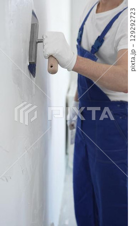 Professional construction worker wearing blue work overall is using a finishing trowel to carefully apply plaster on a wall, demonstrating expertise in home renovation and interior finishing Professional construction worker wearing blue work overall is using a finishing trowel to carefully apply plaster on a wall, demonstrating expertise in home renovation and interior finishing 129292723