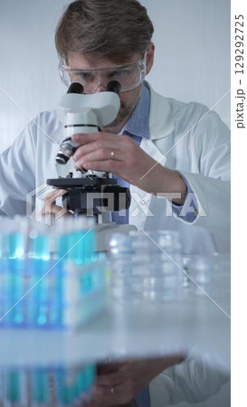 Male scientist researcher wearing lab coat and protective glasses, is using microscope in modern laboratory setup, portrait view. Low key lighting photo. Science concept 129292725