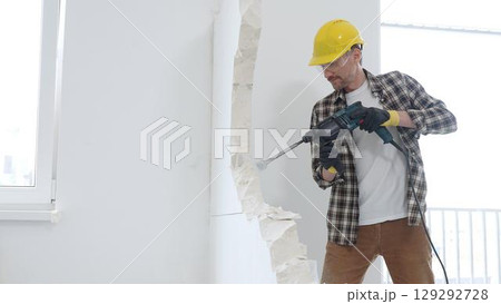 Male construction worker wearing beige checkered shirt, yellow hard hat and protective gloves, is demolishing white wall with rotary hammer drill, generating dust, medium long shot. Renovation project Male construction worker wearing beige checkered shirt, yellow hard hat and protective gloves, is demolishing white wall with rotary hammer drill, generating dust, medium long shot. Renovation project 129292728
