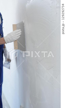 Man construction worker wearing protective gloves and blue construction coveralls, spreading plaster smoothly across wall using professional drywall taping knife Man construction worker wearing protective gloves and blue construction coveralls, spreading plaster smoothly across wall using professional drywall taping knife 129292734