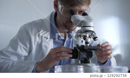 Male scientist wearing lab coat and protective glasses using microscope doing research in modern laboratory setup, portrait view. Low key lighting photo. Science and Medicine concept 129292736