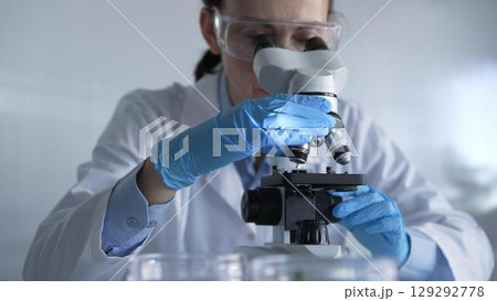 Woman scientist wearing lab coat, white protective gloves and glasses, is using microscope in modern laboratory setup, portrait view. Low key lighting photo. Science and Medicine Woman scientist wearing lab coat, white protective gloves and glasses, is using microscope in modern laboratory setup, portrait view. Low key lighting photo. Science and Medicine 129292778