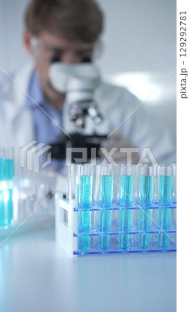 Test tubes with a blue liquid, close-up view. Male scientist wearing white protective gloves and glasses is using microscope on the background. Science and Medicine concept Test tubes with a blue liquid, close-up view. Male scientist wearing white protective gloves and glasses is using microscope on the background. Science and Medicine concept 129292781