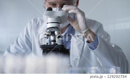 Man scientist researcher wearing white gloves, protective glasses and medical mask is using microscope in modern laboratory, portrait view. Low key lighting photo. Science and Medicine 129292784
