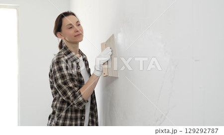 Female construction professional wearing checkered shirt is smoothing white wall surface, applying putty with professional spatula technique, showcasing skilled interior renovation work 129292787