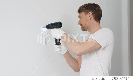 Professional male worker wearing white t-shirt and protective gloves, is drilling hole in a wall with cordless drill during home renovation work. Horizontal portrait view 129292794