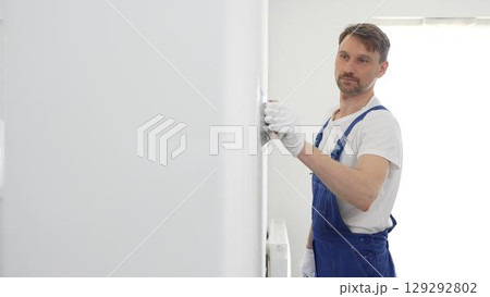 Middle aged male construction worker wearing gloves and blue overalls is using a putty knife to apply plaster on a white wall, performing home renovation 129292802