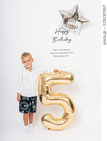 Adorable 5-year-old boy celebrating his birthday with a giant gold number five balloon and festive decor in a bright white setting Adorable 5-year-old boy celebrating his birthday with a giant gold number five balloon and festive decor in a bright white setting 129292997