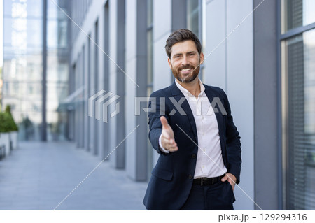 Confident male professional extending a handshake outdoors in a corporate office setting, showcasing business friendliness and a welcoming attitude. 129294316