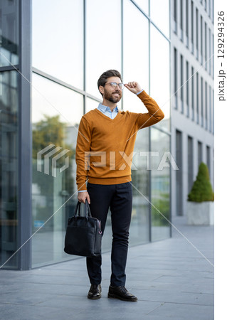 Confidently walking outside a contemporary building, a man wears glasses, formal attire, and carries a black bag, symbolizing a professional and modern lifestyle Confidently walking outside a contemporary building, a man wears glasses, formal attire, and carries a black bag, symbolizing a professional and modern lifestyle 129294326